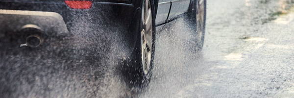 A dark SUV drives away from the camera through puddles of water, splashing the water into the air.